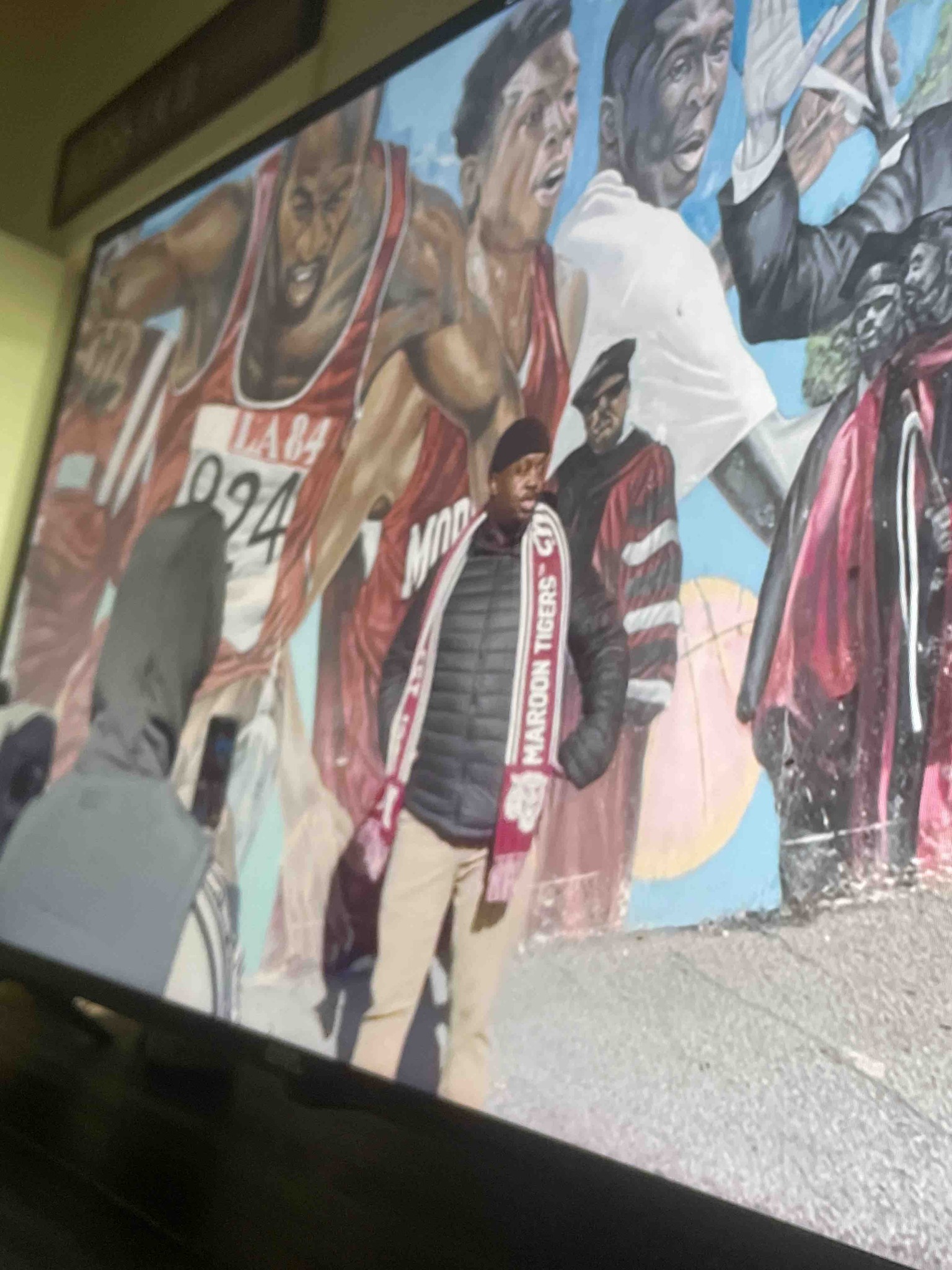 What is the message on the scarf worn by the person in the foreground, and what is the significance of the mural behind him?